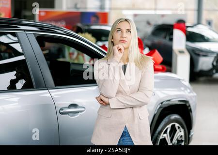 Donna d'affari penosa che sceglie una nuova auto in concessionaria, tocca il mento e guarda in alto, prende la decisione di acquistare un'automobile Foto Stock