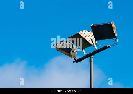 Su un palo alto sono montati tre apparecchi di illuminazione distanziati uniformemente contro un cielo blu vibrante. I dispositivi di fissaggio sono angolati verso l'alto, illuminando l'area sottostante mentre le nuvole si spostano in lontananza. Foto Stock