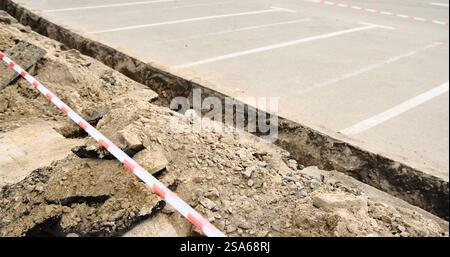 Una trincea appena scavata lungo un parcheggio, contrassegnata con nastro di sicurezza a strisce rosse e bianche. La scena mette in evidenza i lavori di costruzione attivi e la sicurezza Foto Stock