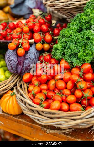 I pomodori ciliegini freschi maturati in vite sono esposti nel cestino di vimini insieme ai cavoli ricci e al cavolo viola allo stand del mercato. Foto Stock