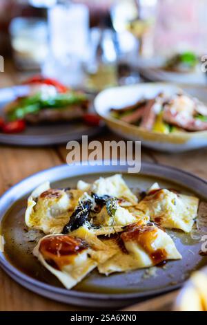 Un piatto di ravioli appena preparati conditi con parmigiano, erbe aromatiche e una salsa leggera, serviti su un tavolo rustico, con altri piatti elegantemente placcati Foto Stock
