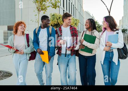 Gruppo di studenti diversi che camminano e parlano con gioia insieme mentre si impegnano in una conversazione vivace e condividono materiali accademici presso il campus universitario pieni di amicizia e Risate. Foto di alta qualità Foto Stock