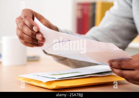 Primo piano delle mani di un uomo nero che mette una lettera all'interno di una busta su una scrivania a casa o in ufficio Foto Stock