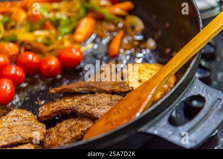 bistecca con cipolle, pomodori e peperoni preparati in una padella su un piano cottura a gas Foto Stock