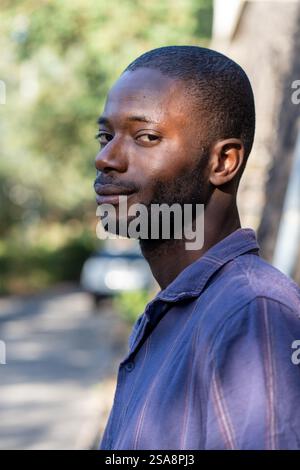 Ritratto ravvicinato di un giovane sicuro di sé con pelle scura che guarda le spalle con un sorriso sottile. L'ambiente naturale all'aperto esalta il suo carisma Foto Stock