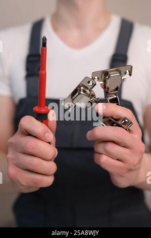 L'assemblatore di mobili tiene in mano una cerniera per mobili e un cacciavite, primo piano. Concetto di costruzione, tuttofare Foto Stock