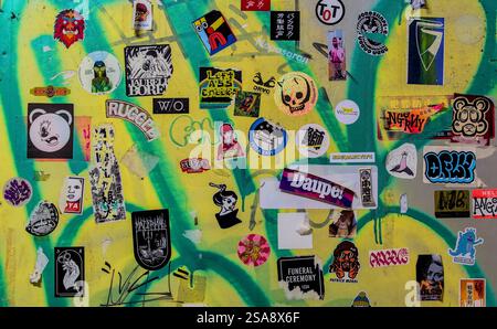 Vista ravvicinata di un muro coperto di graffiti verde e giallo ricoperto da un'eclettica varietà di adesivi Foto Stock