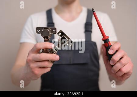L'assemblatore di mobili tiene in mano una cerniera per mobili e un cacciavite, primo piano. Concetto di costruzione, tuttofare Foto Stock