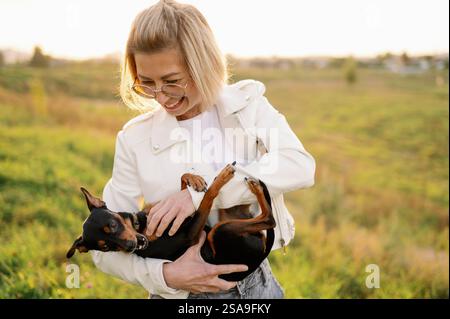 Una donna con gli occhiali tiene un terrier giocattolo russo dai capelli lisci tra le braccia mentre cammina, accarezzandolo. Concetto di amicizia tra uomo e cane, amore f Foto Stock