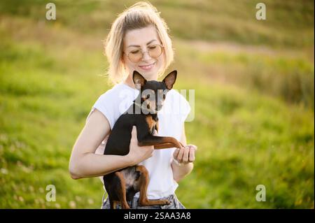Una donna con gli occhiali tiene e abbraccia un giocattolo terrier russo dai capelli lisci mentre cammina in estate. Concetto di amicizia tra uomo e cane, c Foto Stock