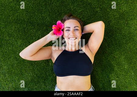 Una donna si sta sdraiando sull'erba con un fiore rosa nei capelli. Sta sorridendo e si sta godendo il momento Foto Stock
