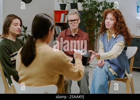 Gruppo di tre persone diverse sedute in un ufficio moderno a discutere di documenti consegnati da una persona. Scaffali e piante sullo sfondo Foto Stock