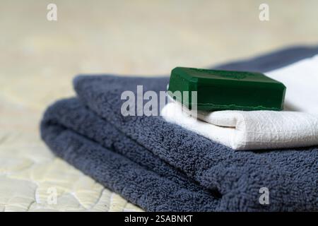 Una pila di asciugamani ben piegati nei colori grigio scuro e bianco, con una barra verde di sapone posta sopra. Lo sfondo presenta un morbido trapuntato sfocato Foto Stock