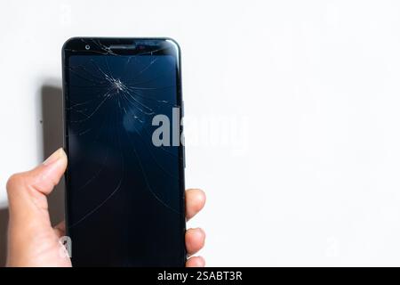 Primo piano di una mano che tiene in mano uno smartphone con uno schermo incrinato su uno sfondo bianco. Lo schermo è scuro e mette in evidenza il tipo ragnatela Foto Stock