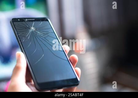 Primo piano di una mano che tiene in mano uno smartphone con uno schermo incrinato su uno sfondo bianco. Lo schermo è scuro e mette in evidenza il tipo ragnatela Foto Stock