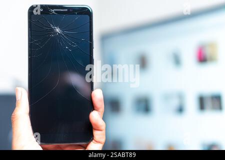 Primo piano di una mano che tiene in mano uno smartphone con uno schermo incrinato su uno sfondo bianco. Lo schermo è scuro e mette in evidenza il tipo ragnatela Foto Stock