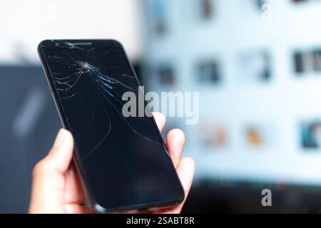 Primo piano di una mano che tiene in mano uno smartphone con uno schermo incrinato su uno sfondo bianco. Lo schermo è scuro e mette in evidenza il tipo ragnatela Foto Stock