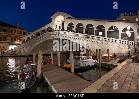 Il Ponte di Rialto di notte nella città di Venezia. Ponte pedonale ad arco in pietra dal 1591 attraverso il Canal grande e gondole ormeggiate su un molo. Foto Stock