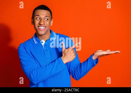 Il giovane sorridente in maglia blu punta in uno spazio vuoto su sfondo arancione Foto Stock