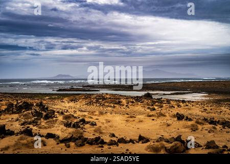 Un suggestivo paesaggio costiero caratterizzato da terreno roccioso, aree sabbiose e turbolente onde oceaniche sotto un cielo nuvoloso. Montagne lontane sono visibili sul Foto Stock