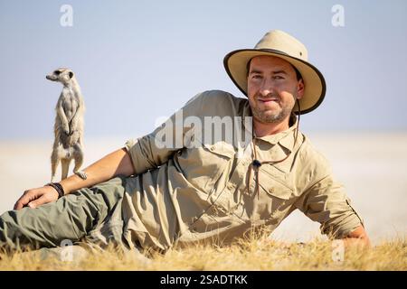Un uomo in abbigliamento da safari, sdraiato su erba asciutta sotto il sole, accompagnato da un meerkat in piedi alermente accanto a lui, si trova su un tranquillo dorso naturale Foto Stock