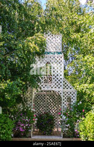 Un gazebo bianco con tetto a reticolo si trova in un lussureggiante giardino verde. Il gazebo e' circondato da una varieta' di fiori e piante, creando un sereno e pisello Foto Stock