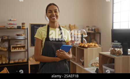 Donna sorridente all'interno di una panetteria che tiene in mano un tablet, circondata da vari dolci in mostra, creando un'atmosfera calda e invitante. Foto Stock