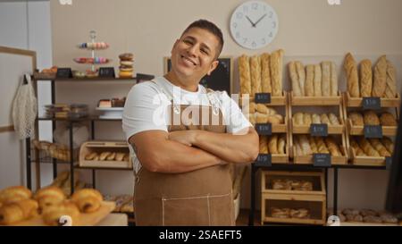 Sorridente giovane ispanico con le braccia incrociate in una panetteria, circondato da pane e dolci freschi, in piedi con fiducia in un negozio latino al coperto Foto Stock