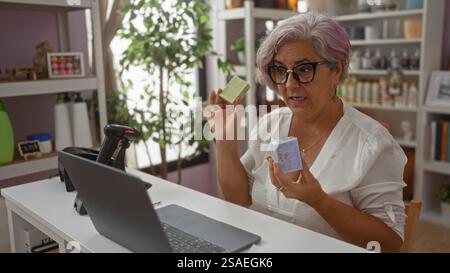 Una donna matura con i capelli grigi mostra saponette durante una videochiamata dal suo ufficio su uno sfondo rosa Foto Stock