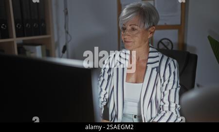 Donna dai capelli grigi corti e un blazer a righe che lavora su un computer desktop in un ufficio moderno, trasmettendo un professionista e concentrandosi su Foto Stock