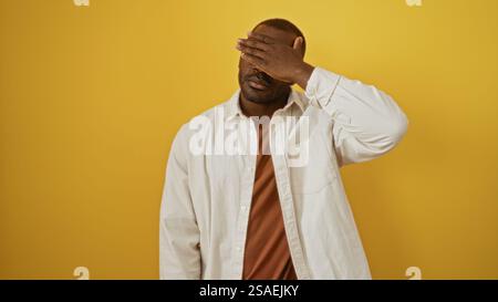 Giovane, nero, uomo che si copre il viso con una mano isolata su un vibrante sfondo giallo, indossa una camicia bianca ed esprime emozioni Foto Stock