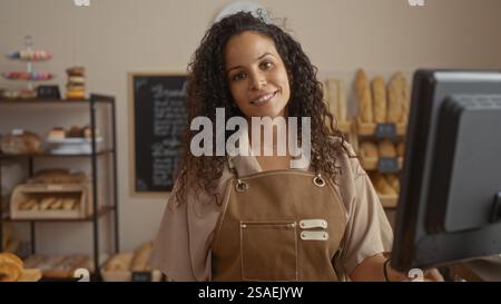 Donna che lavora in una panetteria con pane e pasticcini sullo sfondo, mostrando una giovane, bella, bruna e ispanica che sorride alla macchina fotografica Foto Stock