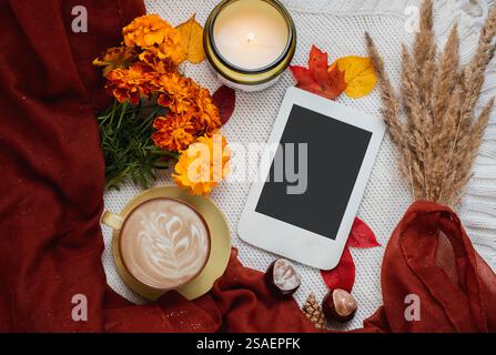 Accogliente composizione autunnale con sciarpa Bordeaux, tazza di caffè, candela accesa, foglie autunnali ed e-book con spazio per le copie. Foto Stock