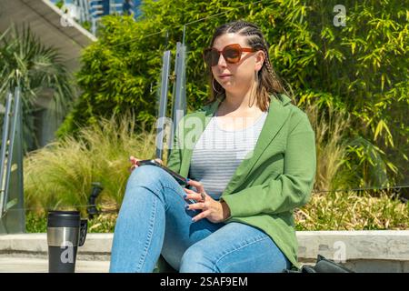 Una donna rilassata si gode una pausa all'aperto, tenendo una tazza di caffè e controllando il suo smartphone in una giornata di sole. Perfetto per concetti di svago, tecnologia, Foto Stock