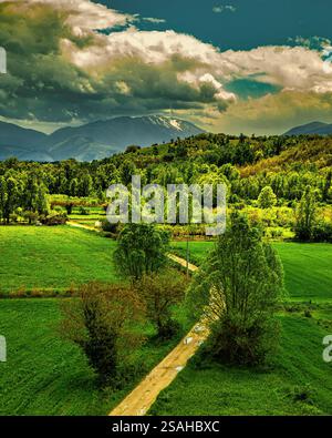 Paesaggio panoramico che mostra campi verdi attraversati da una tortuosa strada sterrata che conduce a montagne innevate sotto un cielo nuvoloso. Abruzzo, Italia, Europa Foto Stock