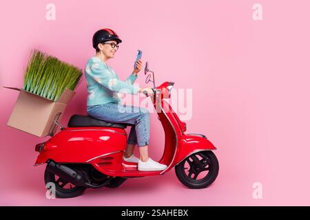 Donna anziana in sella a uno scooter rosso con piante su sfondo rosa mentre utilizza lo smartphone Foto Stock