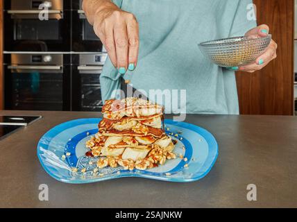 Una persona cosparge le noci tritate su una pila di pancake a strati di banane e sciroppo, finendo il piatto della colazione. Foto Stock