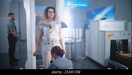 Terminal dell'aeroporto: Donna attraversa i cancelli del rilevatore di metalli, porta i bagagli dal nastro trasportatore. L'agente di sicurezza controlla i passeggeri e i documenti per il volo aereo. Diversi turisti che vanno in vacanza. Foto Stock