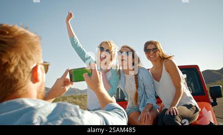 Giovane uomo con telefono cellulare che scatta foto di tre donne durante il viaggio su strada seduto sul cofano dell'auto Foto Stock