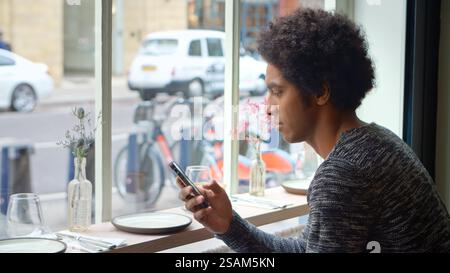 Giovane seduto al finestrino del Cafe per controllare i messaggi o i social media utilizzando il telefono cellulare Foto Stock