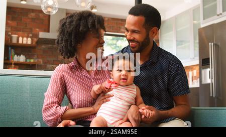 Famiglia sorridente seduta sul divano a casa che tiene in mano la bambina Foto Stock