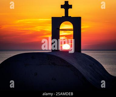 Una splendida vista del tramonto incorniciata da una struttura architettonica bianca con una croce. Il sole tramonta sull'oceano, emettendo calde tonalità arancio e giallo Foto Stock