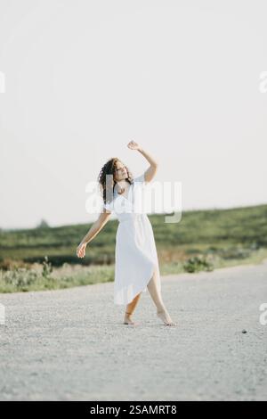 Donna in abito bianco con le braccia alzate, ballando su un sentiero immerso nel verde Foto Stock