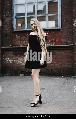 Donna che posa con un vestito nero contro un muro di mattoni con finestre Foto Stock