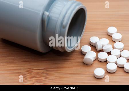 Una bottiglia di plastica grigia, probabilmente un contenitore per farmaci, è aperta su una superficie di legno marrone chiaro. Le pillole bianche e rotonde sono sparse intorno alla bottiglia Foto Stock