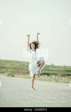 Donna che balla allegramente con le braccia alzate con un abito bianco in una giornata di sole Foto Stock