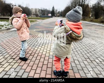 Due bambini che si divertono a fotografare in una giornata fredda all'aperto, mostrando creatività giovanile e momenti divertenti con le telecamere in un tranquillo parco urbano. Foto Stock