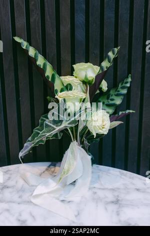 Una disposizione floreale splendidamente realizzata presenta rose verdi e foglie vivaci, elegantemente esposte su un tavolo di marmo su uno sfondo scuro e foderato Foto Stock
