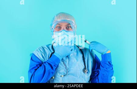 Medico uomo in maschera medica, cappuccio chirurgico e guanti con siringa. Medico o infermiere di sesso maschile in tuta protettiva e occhiali con stetoscopio che regge syri Foto Stock