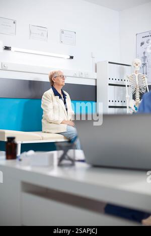 Donna anziana vestita in modo informale siede pazientemente guardando le immagini sul muro dello studio medico, pronto per l'appuntamento dei medici. Donna anziana caucasica aspetta sul letto d'ospedale, preparata per l'esame di controllo. Foto Stock
